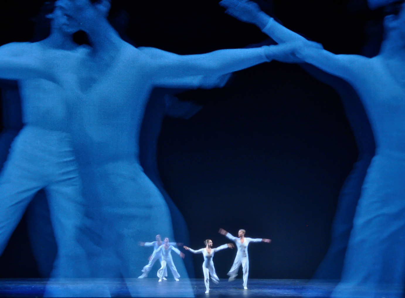 Four dancers cavort in front of dancers on film. Everyone is dressed in white pants and long-sleeved tops.
